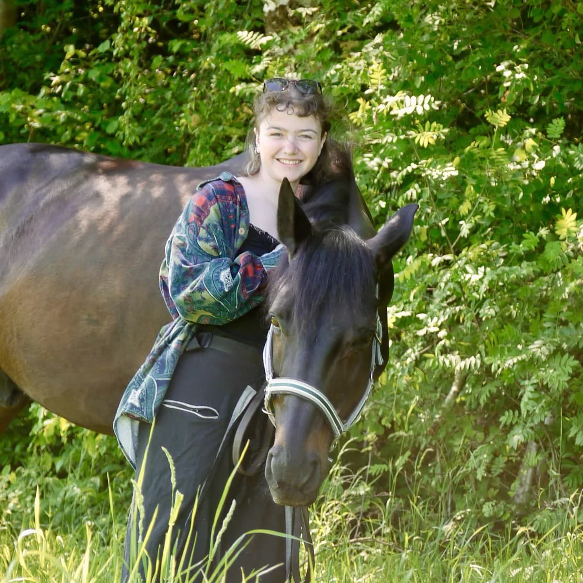 Fiete steht lächelnd bei ihrem dunkelbraunen Pferd auf einer sommerlichen Wiese